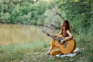 un hippie mujer jugando su guitarra sonrisas y canta canciones en naturaleza sentado en un tartán en el noche en el puesta de sol luz de sol. un estilo de vida en armonía con el cuerpo y naturaleza foto