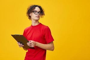 Confused myopic young student man in red t-shirt funny eyewear holds tablet folder with study notes looks aside posing isolated on over yellow background. Free place for ad. Education College concept photo