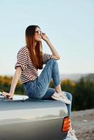 A fashion woman in stylish sunglasses, a striped t-shirt and jeans sits on the trunk of a car and looks at the beautiful nature of autumn. Travel lifestyle photo