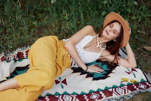 A woman in eco-friendly clothing in a hippie look is lying on a colored plaid smiling and looking at an autumn sunset in nature in the park. Lifestyle on a journey of harmony with the world photo