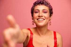 Moda retrato de un mujer con un corto Corte de pelo de púrpura color y un sonrisa con dientes en un rojo parte superior en un rosado antecedentes bailando felizmente foto