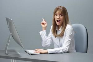Excited happy blonde businesswoman freelancer have cool idea point finger up using desktop computer sitting at workplace in gray modern office. Remote Job And Career Profession Concept. Copy space photo