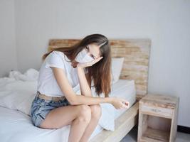 sad woman in medical mask sitting on the bed with her head tilted down pandemic isolation model photo