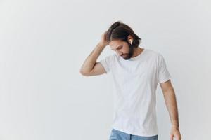 A man with a beard and long hair in a white T-shirt and blue jeans stands against a white wall, leaning against it and listening to music with wireless white headphones, staring thoughtfully photo