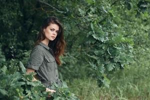 mujer en el bosque mantiene su manos en su bolsillos con verde hojas antecedentes foto