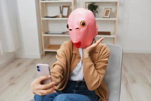 Happy funny blonde lady in pink fish mask warm sweater doing selfie video call sitting in armchair at modern home interior. Pause from work, take a break, social media in free time concept. Copy space photo