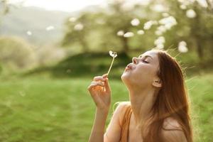 The happy woman smiles and blows the dandelion in the wind. Summer green landscape and sunshine in the background photo