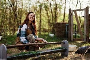 un hembra pájaro granja trabajador sonrisas y es contento torrencial comida dentro el pollo alimentador en el Fresco aire sentado en el verde césped foto