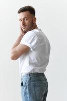 View from the back shot of handsome serious tanned man guy in basic t-shirt looks at camera posing on white background. Fashion Style New Collection Offer. Copy space for ad. Model snap photo