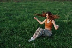 un Pelirrojo mujer con largo, azotado por el viento pelo se sienta al aire libre en el césped en el parque y sonrisas, el puesta de sol ligero esclarecedor su rostro. el concepto de armonía con naturaleza al aire libre foto