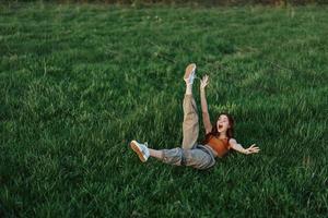 A young woman playing games in the park on the green grass spreading her arms and legs in different directions falling and smiling in the summer sunlight photo