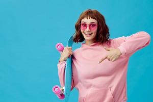 Funny joyful pretty redhead lady in pink hoodie sunglasses with penny board smiling posing isolated on blue studio background. Copy space Banner Offer. Fashion Cinema. Holiday activity photo