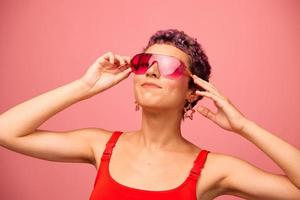 Moda retrato de un mujer con un corto Corte de pelo en de colores Gafas de sol con raro accesorios con pendientes sonriente en un rosado brillante antecedentes con un aptitud cuerpo bailando foto