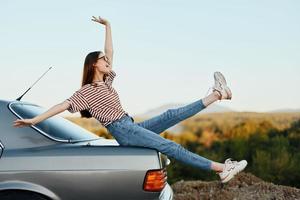 un mujer con un coche detenido en el la carretera a descanso en el viaje elevado su brazos y piernas desde felicidad y un hermosa paisaje foto