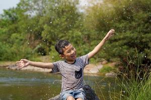 Asian boy taking a break from playing in a stream Sit on the rock, smiling brightly, arms outstretched to enjoy the splashing water happily. soft and selective focus. photo