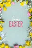 Happy Easter text with spring flowers flat lay frame composition on colored background. Vertical Easter greeting card photo