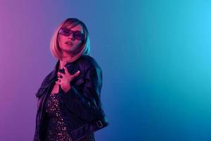 Pretty smiling sexy beautiful blonde woman touch leather jacket sparkly dress trendy sunglasses posing isolated in blue pink color light studio background. Neon party Cyberpunk concept. Copy space photo