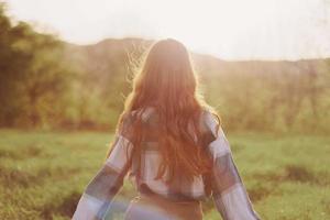 un mujer carreras a través de un campo con su espalda a el cámara en un verano día con su pelo largo y volador en el puesta de sol. el concepto de libertad y armonía con naturaleza foto