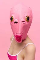 un mujer vistiendo un silicona Víspera de Todos los Santos máscara en el forma de un rosado pescado con grande amarillo ojos mira a el cámara en contra un rosado antecedentes foto