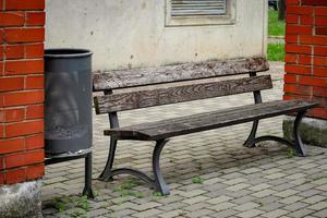 Brown wooden bench with black metal legs standing in a yard between two building columns of red brick near a black trash bin photo