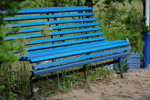 azul de madera banco con metal piernas en pie en un parque con descuidado verde arbustos y arena la carretera un azul pintado basura compartimiento con basura en y fuera foto