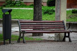 Brown wooden bench with a decorative ornate metal legs standing in a park with green lawn near a black trash bin photo