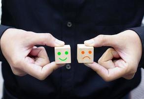 Businessman holding smiling and non-smiling face icons which print screen on wooden cube block. Emotion, mindset and satisfaction concept. photo