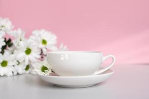 A white cup with a plate stands on a pink background with white chrysanthemum flowers photo