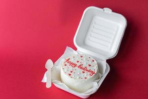 festivo bento cumpleaños pastel con corazones en un caja con un de madera cuchara. rojo antecedentes con cintas foto