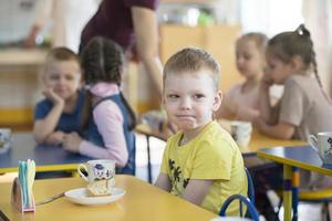 niños comer en jardín de infancia. chico con un taza a el mesa en el antecedentes de niños foto