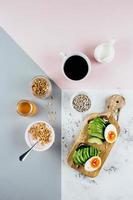 Sandwich with avocado, boiled eggs, yogurt with granola, cup of coffee over tricolor background. photo