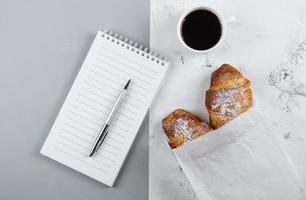 Coffee mug with croissants, empty notebook, pen on two-color background from above. Morning routine photo