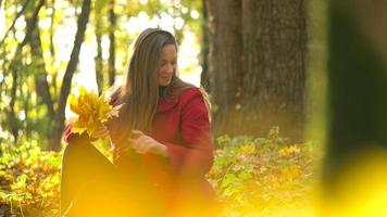 Portrait of a beautiful smiling girl with a yellow maple leaf in the foreground in the autumn forest. Slow motion video