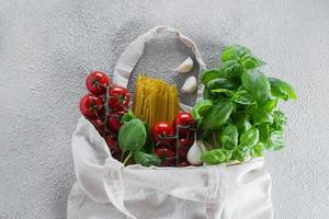 Reusable bag with groceries. Minimal waste. Basil, tomatoes cherry, garlic in fabric bag. Top view, photo