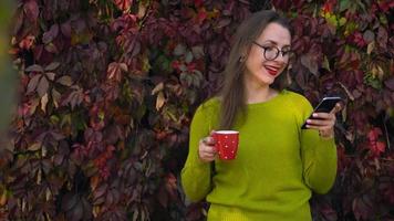 mujer con lentes bebidas té o café al aire libre y usos un teléfono inteligente en un calentar otoño día. café descanso video