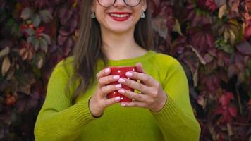 kvinna med glasögon drycker te eller kaffe utomhus och användningar en smartphone på en värma höst dag. kaffe ha sönder video