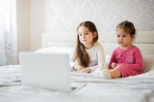 Two cute little sisters wear pajamas looking cartoon by laptop in morning at home. photo