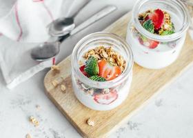 Two jars with tasty parfaits made of granola, strawberries and yogurt on white marble table. photo