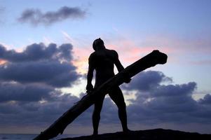 un muscular hombre levantamiento arriba un grande rama o Iniciar sesión a el playa en un dramático silueta posición. foto