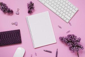 Women's desktop close-up, keyboard, notebooks with pen, lilac flowers. Minimal cropped flat composition on a pink background. Copy space photo