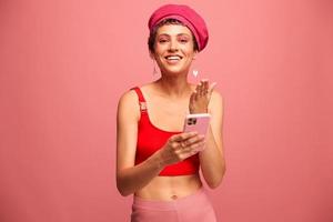 Young woman blogger with colored pink hair and short haircut flipping through the phone screen and typing a message with a smile and surprise in stylish clothes on a pink background photo