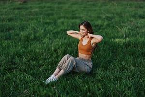 A young redheaded woman sits on the green grass in the park with her arms outstretched in different directions and smiling in the light of the setting sun photo