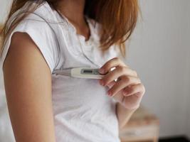 A woman in a white t-shirt is checking the temperature with a thermometer under her arm photo