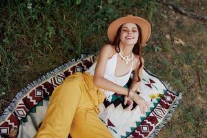 A woman in eco-friendly clothing in a hippie look is lying on a colored plaid smiling and looking at an autumn sunset in nature in the park. Lifestyle on a journey of harmony with the world photo