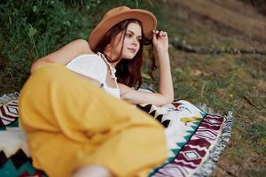 A woman in eco-friendly clothing in a hippie look is lying on a colored plaid smiling and looking at an autumn sunset in nature in the park. Lifestyle on a journey of harmony with the world photo