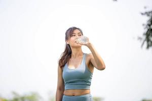 Athlete young beautiful woman drinking water from a plastic bottle at summer green park, Sport woman drinking water after work out exercising photo