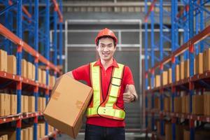 Male worker in hardhat holding cardboard box walking through in retail warehouse, Warehouse worker working in factory warehouse, Man carrying box and showing thumbs up photo