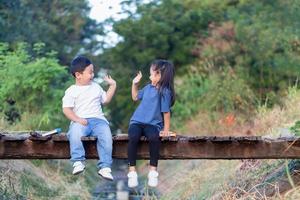 alegre niños sentado en de madera puente, asiático niños jugando en jardín, chico y niña leyendo libros foto