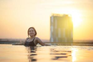 silueta de asiático mujer relajante en infinito nadando piscina con puesta de sol ver con alto subir skyscape urbano centro, felicidad estilo de vida foto