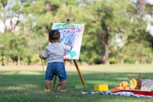 espalda ver de niño chico pintura en lona en el jardín, contento niño chico dibujo un imagen al aire libre foto